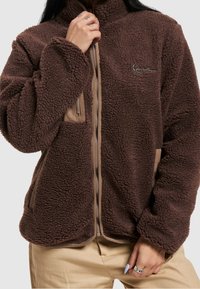 Giacca in pile marrone con colletto alto, apertura con zip, dettagli beige e tasche laterali. Presenta una superficie testurizzata e soffice.