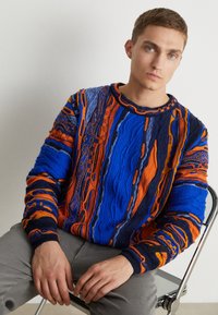 Strickpullover in lebhaftem Blau und Orange, mit strukturierten Mustern und gerippten Bündchen. Modell sitzt und trägt graue Hosen.