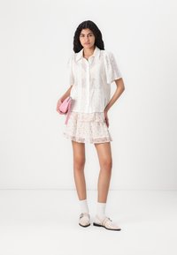 Blusa bianca in pizzo con maniche a palloncino, gonna trasparente a strati con fiori, scarpe crema e borsa rosa, su uno sfondo bianco uniforme.