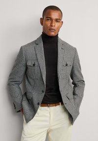 Checkered blazer in black and white, double-breasted style with two front pockets, worn over a fitted black turtleneck. Light-colored trousers.