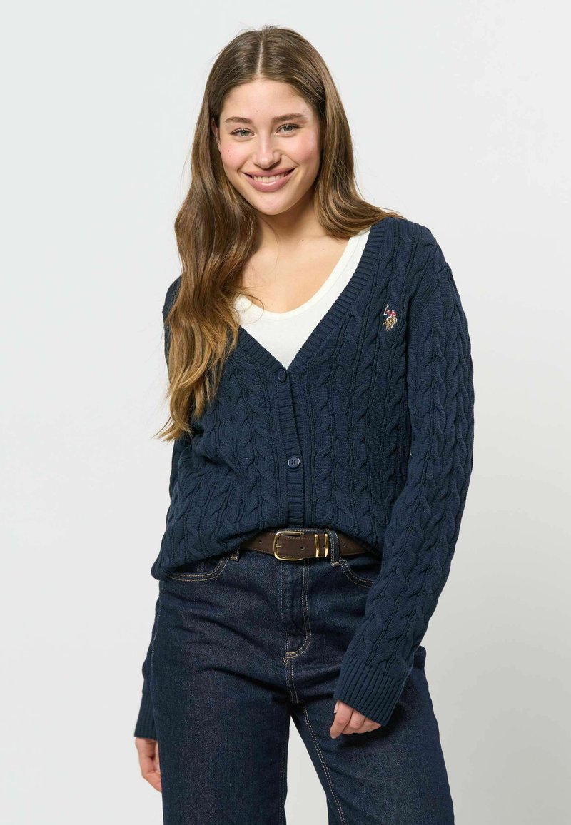 Jeune femme portant un cardigan bleu marine torsadé sur un top blanc avec un jean foncé et une ceinture marron, souriante à la caméra.