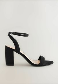 Sandale à talon haut en daim noir avec un talon carré, une sangle de cheville réglable, un design à bout ouvert et une semelle plate.