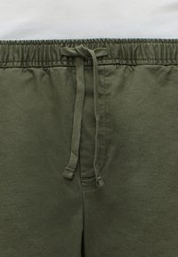 Gros plan sur un pantalon vert olive avec une taille élastique et un cordon noué à l’avant sur fond blanc.