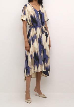 Kobieta ubrana w sukienkę na guziki w niebieskie, beżowe i czarne wzory, z krótkimi rękawami i wiązanym paskiem, zestawioną z beżowymi mulesami na obcasie z zaostrzonym noskiem.