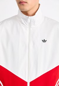 White and red zip-up jacket featuring a high collar, textured fabric, and a small black logo. The design includes bold color blocking.