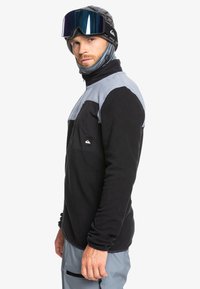 Czarny i szary polarowy żakiet z półzamkiem, kieszenią na piersi i logo Quiksilver. Charakteryzuje się kontrastującymi górnymi częściami oraz miękką teksturą.