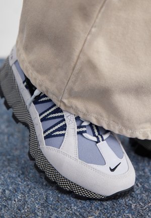 Gros plan d’un pied portant un pantalon beige et une basket Nike blanche, bleue et noire avec une semelle à motifs sur un sol texturé.