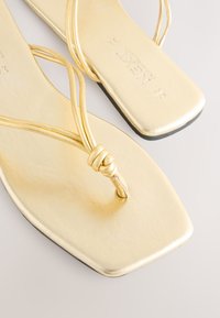 Goldene flache Sandalen mit dünnen Riemen, einem Knoten-Detail und einer quadratischen Zehenpartie. Das metallische Finish verleiht Glanz, während die Sohle schwarz ist.