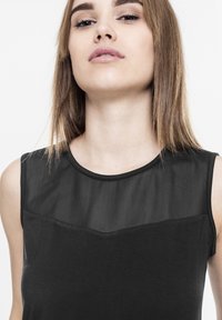 Ung kvinde med lyst brunt, glat hår iført en ærmeløs sort top med en gennemsigtig mesh-udskæring mod en hvid baggrund.