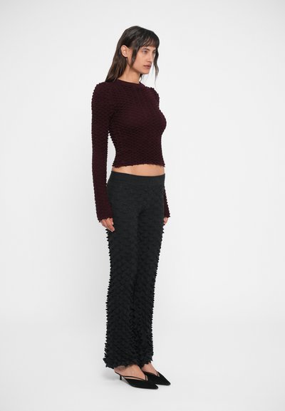 Burgundi textúrájú, hosszú ujjú blúz fekete fodros nadrággal párosítva. Az anyag puhanak tűnik, réteges részletekkel és slim fit fazonnal. Fekete, hegyes orrú cipők.