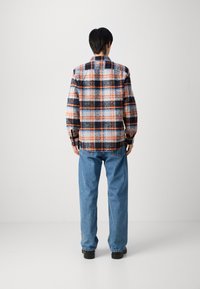 Giacca in flanella a quadri blu, arancione e nera; vestibilità comoda con maniche lunghe, polsini a bottone e dettaglio del colletto sul retro. Abbinata a jeans di denim larghi.