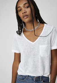 White V-neck t-shirt in lightweight, semi-sheer fabric with a front pocket and rolled sleeves, paired with blue denim jeans.