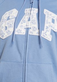 Sweat à capuche zippé bleu clair en matière douce, avec de grandes lettres en appliqué floral "GAP" et des cordons de serrage sur la capuche.