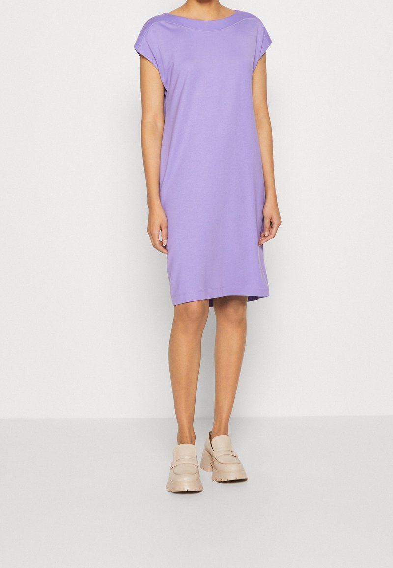 Femme debout devant un fond blanc uni, portant une robe ample à manches courtes lavande et des mocassins beiges épais. Visage non visible.