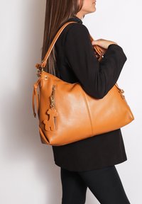 Donna con cappotto nero che tiene una grande borsa di pelle beige con un portachiavi a forma di orsetto su sfondo bianco.