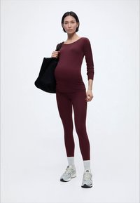 Raseeritud naine, kes kannab merevaigu toonis tihedaltistuvat pika varrukaga t-särki ja retuuse, valgeid sokke, hallid tossud, kandes musta suurt kandekotti õlal.