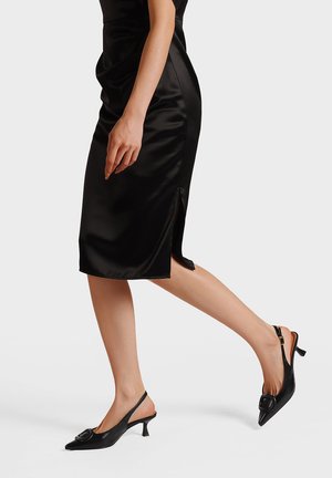 Vestito in raso nero con design aderente e spacco laterale, abbinato a décolleté slingback nere con punta affilata e dettaglio a fiocco.