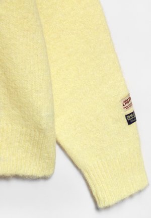 Zachte gele gebreide trui-mouw met geribde manchet en twee kleine stoffen labels dichtbij de pols.