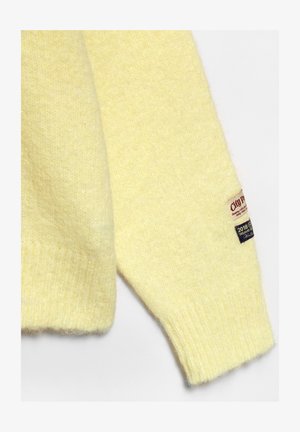 Blød gul strikket sweaterærme med ribbet manchet og to små stofmærker nær håndleddet.