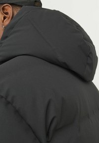 Paminkštintas juodas švarkas su aukštu apykakle ir pritvirtinta kapišonu. Pasižymi lygiu išoriniu paviršiumi ir minimalistiniu dizainu su ergonomiškomis siūlėmis.