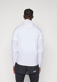 Giacca leggera bianca con maniche lunghe e cappuccio aderente. Tessuto morbido, design minimalista, abbinata a pantaloni corti scuri con logo.