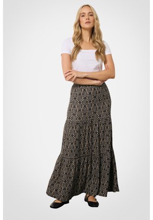 Vrouw met lang blond haar draagt een wit topje met korte mouwen, een zwarte maxi rok met patroon en zwarte platte schoenen, staand met gekruiste armen.