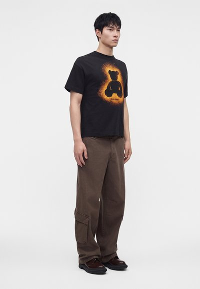 Νέος άνδρας στέκεται φορώντας μαύρο T-shirt με πορτοκαλί φωτεινή εικόνα με αρκουδάκι, καφέ παντελόνια cargo και σκούρα καφέ παπούτσια.