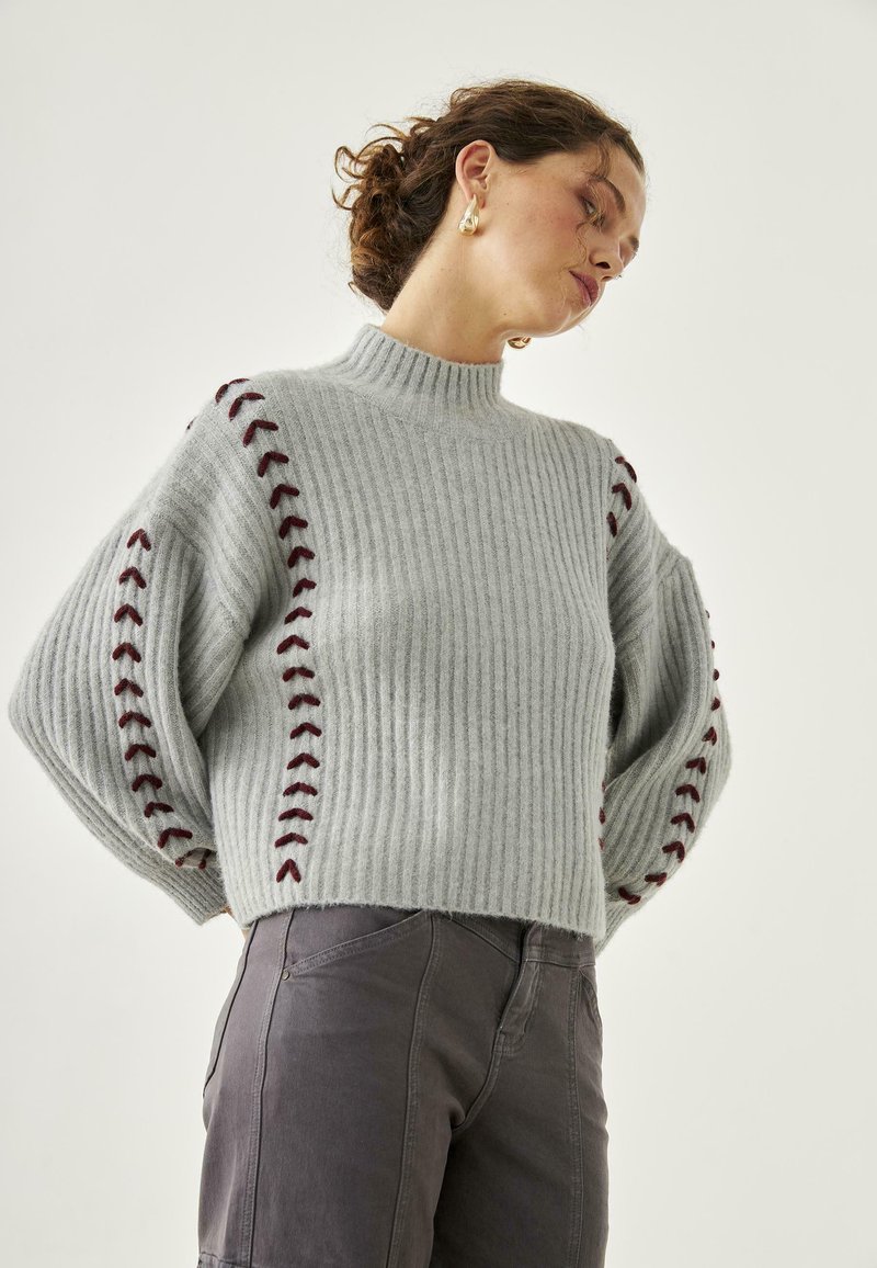 Suéter de canalé gris claro con cuello alto y acentos de costura en burdeos a lo largo de las mangas. Textura suave, ajuste holgado y longitud corta.