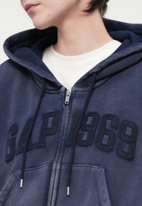 Tamno plava zip-up dukserica s velikim reljefnim natpisom "GAP 1969", sa vezicama i brušenom teksturom. Preko nje je složen bijeli majica s okruglim izrezom.