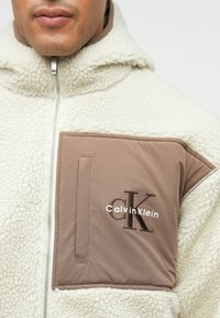 Casaco de fleece em creme com um bolso de nylon castanho no peito. Apresenta um fecho éclair e o logo "Calvin Klein", combinando uma textura suave com materiais duráveis.