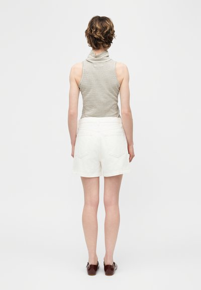 Άτομο που στέκεται με την πλάτη στον θεατή, φοράει αμάνικη ριγέ ζιβάγκο μπλούζα, λευκά σορτς και σκούρα καφέ παπούτσια σε απλό φόντο.