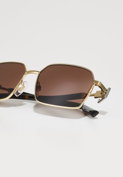 Gold-framed sunglasses with a hexagonal shape, brown tinted lenses, and black temple arms; features a clear nose pad and silver hardware accents.
