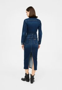 Abito in denim di colore blu scuro, aderente con maniche lunghe, chiusura a bottoni e spacco posteriore; abbinato a stivaletti neri sopra la caviglia. Tessuto testurizzato, lunghezza media.
