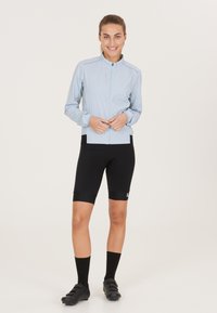 Helle blaue leichte Jacke mit einem Reißverschluss vorne, langen Ärmeln und elastischen Bündchen. Schwarze Radsportshorts und schwarze Schuhe vervollständigen das Outfit.