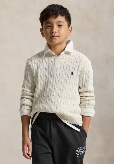 Jeune garçon portant un pull en maille torsadée blanc sur une chemise blanche à col et un pantalon de survêtement foncé avec le logo Polo, debout devant un fond uni.