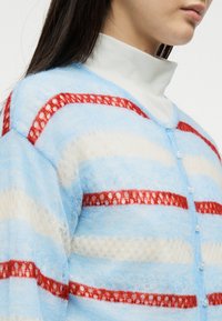 Γυναίκα που φοράει λευκό ζιβάγκο κάτω από μια απαλή, ανοιχτό μπλε ζακέτα με οριζόντιες κόκκινες και άσπρες ρίγες και κούμπωμα με κουμπιά.