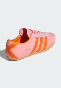 Sneakers roz cu accente portocalii, având trei dungi și o talpă din cauciuc. Materialul pare neted cu o limbă din velur și detalii de branding.