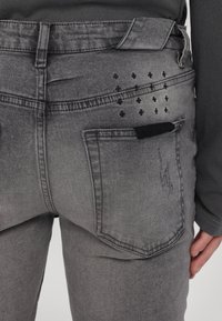 Hallitatud hallid denim teksad, millel on veidi kulunud tekstuur. Tagaküljel on tasku koos õmblusdetailidega ja väike must kangast aksessuaar.