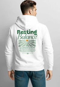 Mann trägt einen weißen Hoodie mit dem Schriftzug "Resting Balance" und einer grünen Grafik zur Förderung der psychischen Gesundheit auf dem Rücken, kombiniert mit blauen Jeans.