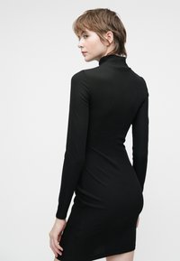 Vestido negro de manga larga con cuello alto, diseño ajustado, textura acanalada y dobladillo a la altura de la rodilla. La parte posterior es lisa con costuras mínimas.