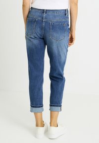 Vaqueros azules de mezclilla con dobladillo enrollado, dos bolsillos traseros y un ligero desgaste. Combinados con zapatillas blancas. Diseño moderno y casual.