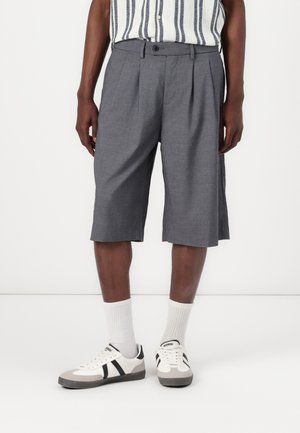 Szare, sięgające do kolan szorty z zakładkami, wykonane z lekkiej tkaniny. Noszone z białymi skarpetkami i sportowymi butami z czarnymi paskami.