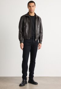 Chaqueta bomber de cuero marrón con cierre de cremallera, puños acanalados y bolsillos laterales, combinada con jeans ajustados negros y botas negras.
