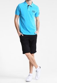 Polo bleu clair avec un col noir, manches courtes et logo sur le devant. Porté avec un short noir et des baskets blanches pour un style décontracté.