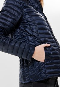 Námorní sky puffer bunda s prošívaným designem, pocházející s stojáčkem a bočními kapsami, vyrobená z lesklého, lehkého materiálu.