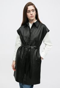 Fekete bőrből készült ballonkabát gombos elülső résszel, rövid ujjakkal, derékrészen övvel és két oldalzsebbel. Simított textúrával és strukturált kialakítással rendelkezik.