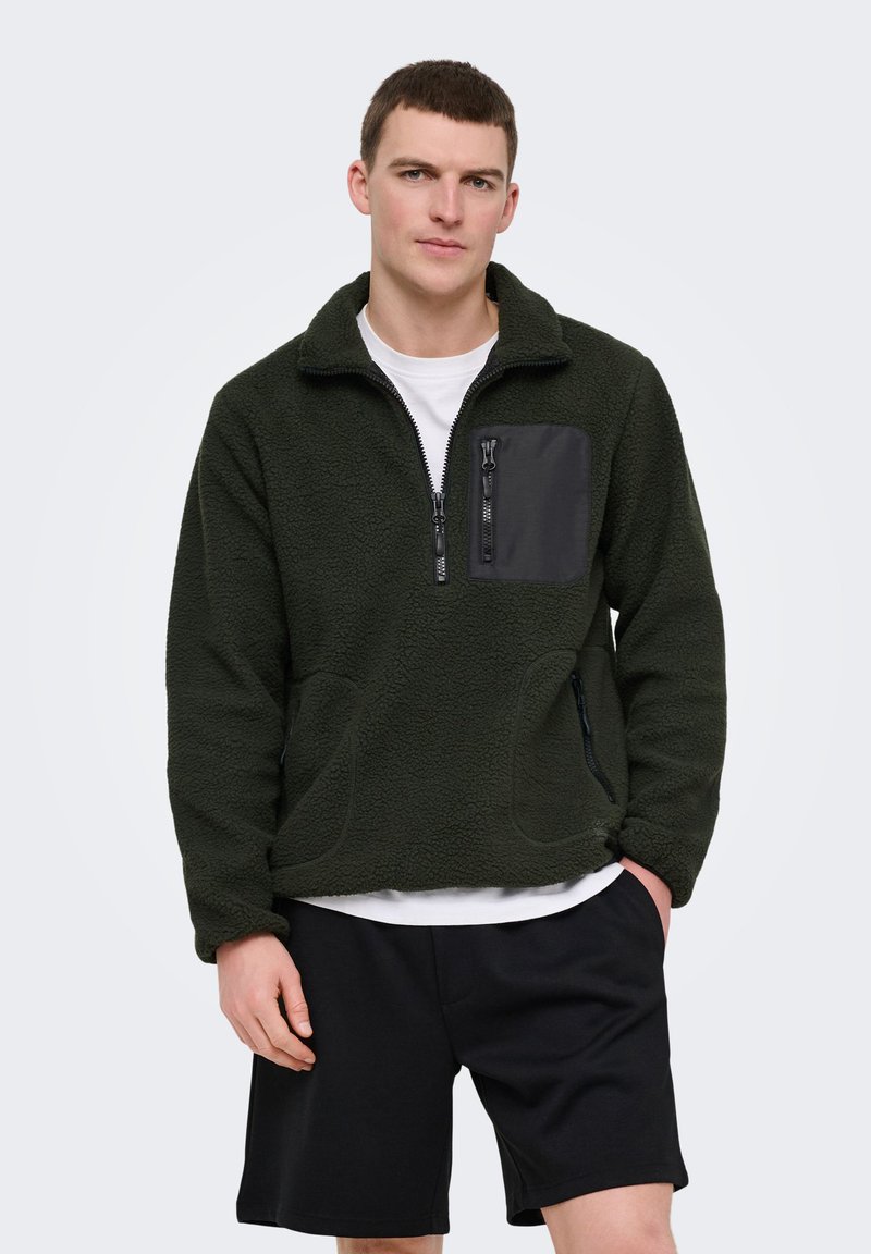 Jeune homme portant une veste polaire vert foncé avec des poches zippées et un short noir, debout avec une main dans la poche devant un fond uni.