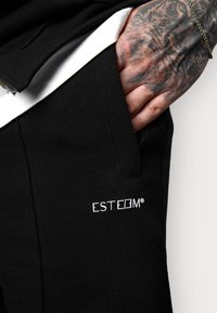 Joggers en coton noirs avec poches latérales, arborant un logo brodé blanc "ESTEEM" sur la cuisse. Vue rapprochée avec un design texturé et décontracté.