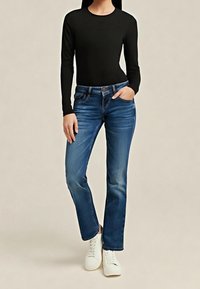 Femme portant un haut noir à manches longues, un jean droit bleu, des baskets blanches, se tenant debout avec une main dans la poche devant un fond uni.