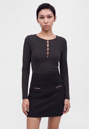 Femme portant un haut noir à manches longues avec des lettres dorées "GUESS" sur le devant et une jupe noire avec des accents de chaînes dorées sur les poches.